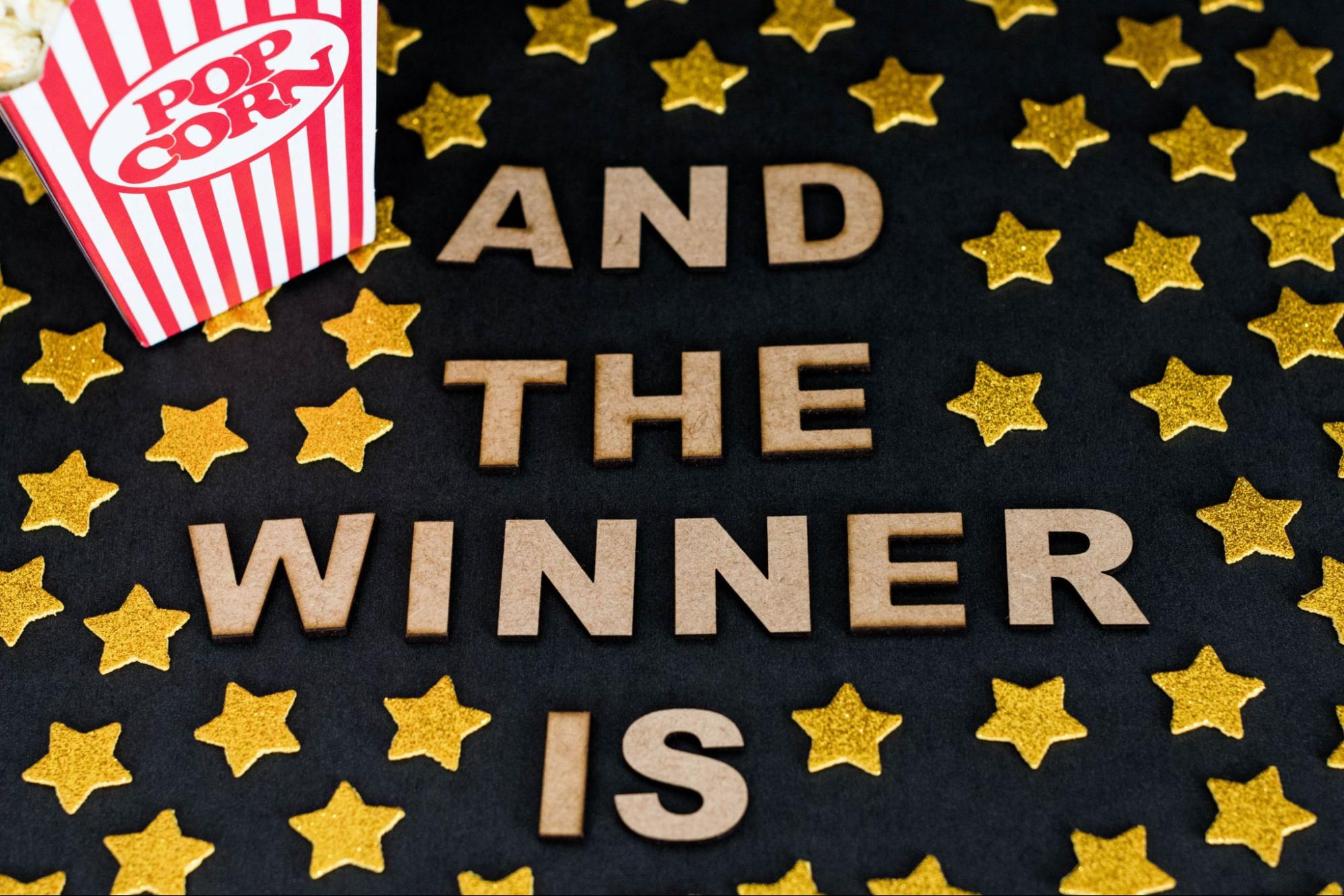 Celebratory 'AND THE WINNER IS' text with popcorn and gold stars on a black background, showcasing famous awards and trophies from DG Trophies, perfect for award events.